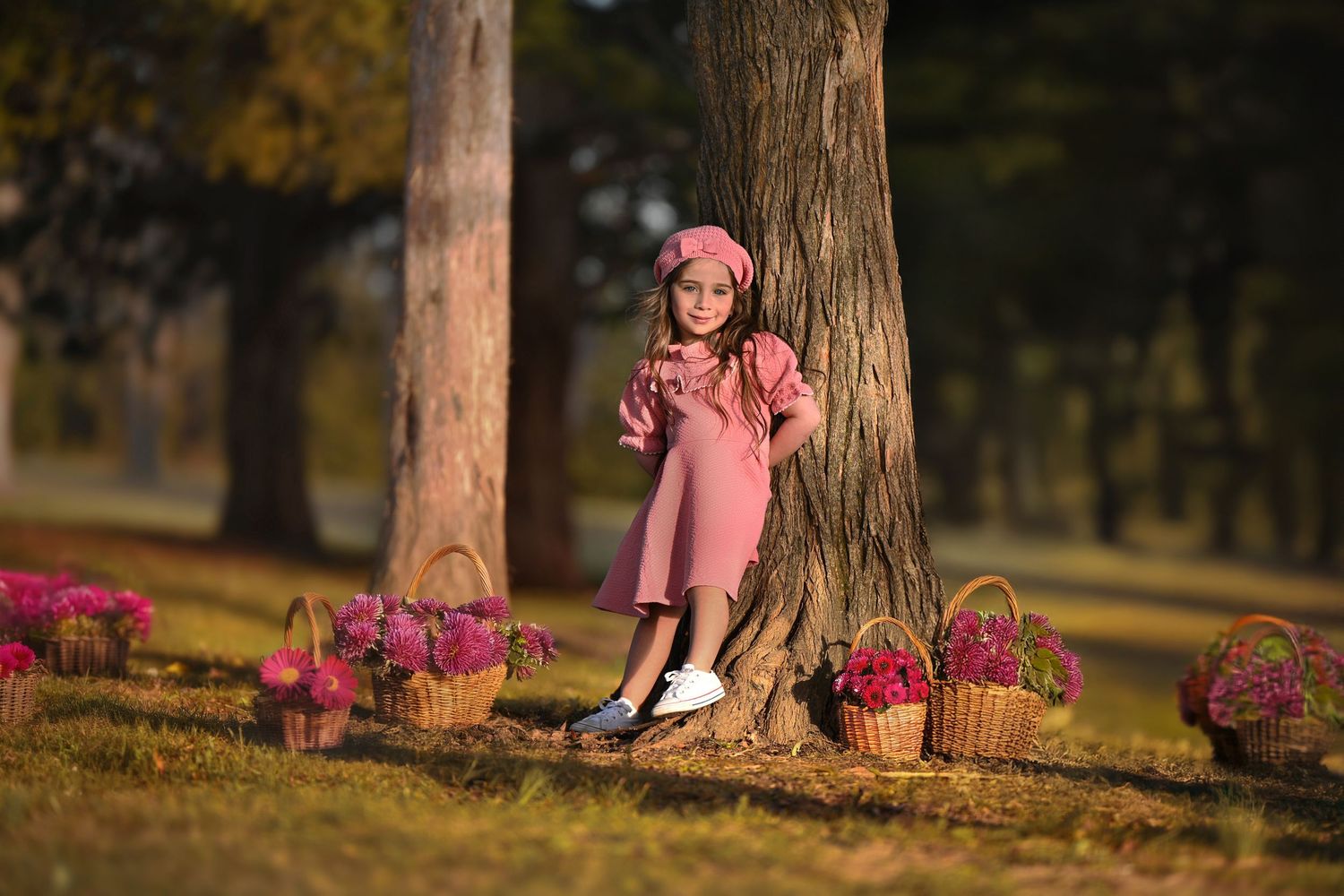 A child in a pink coat stands by a tree trunk surrounded by pink flowers in a sun-dappled autumn park.