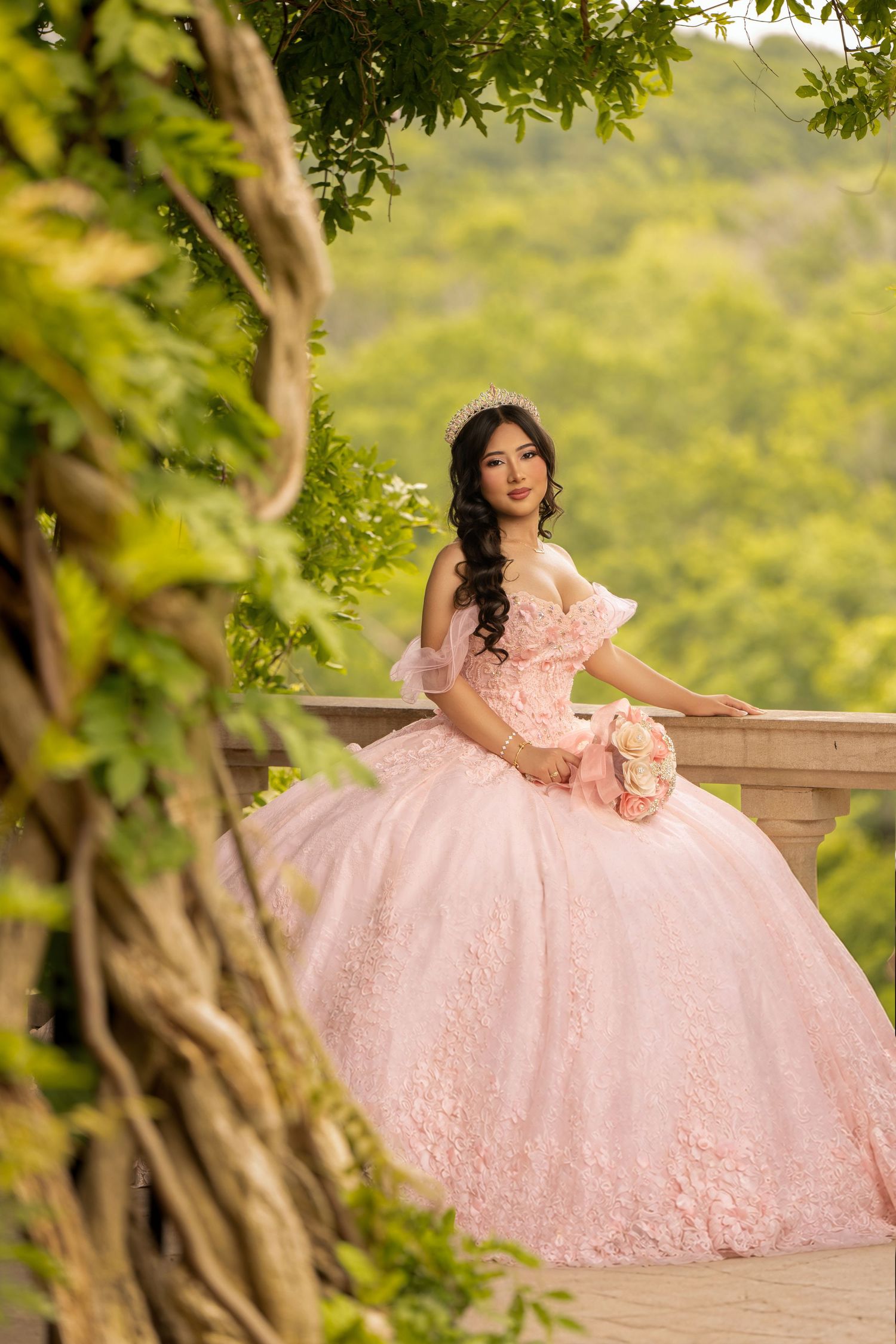 Young woman in pink ball gown with floral details holds bouquet on balcony surrounded by lush green trees.