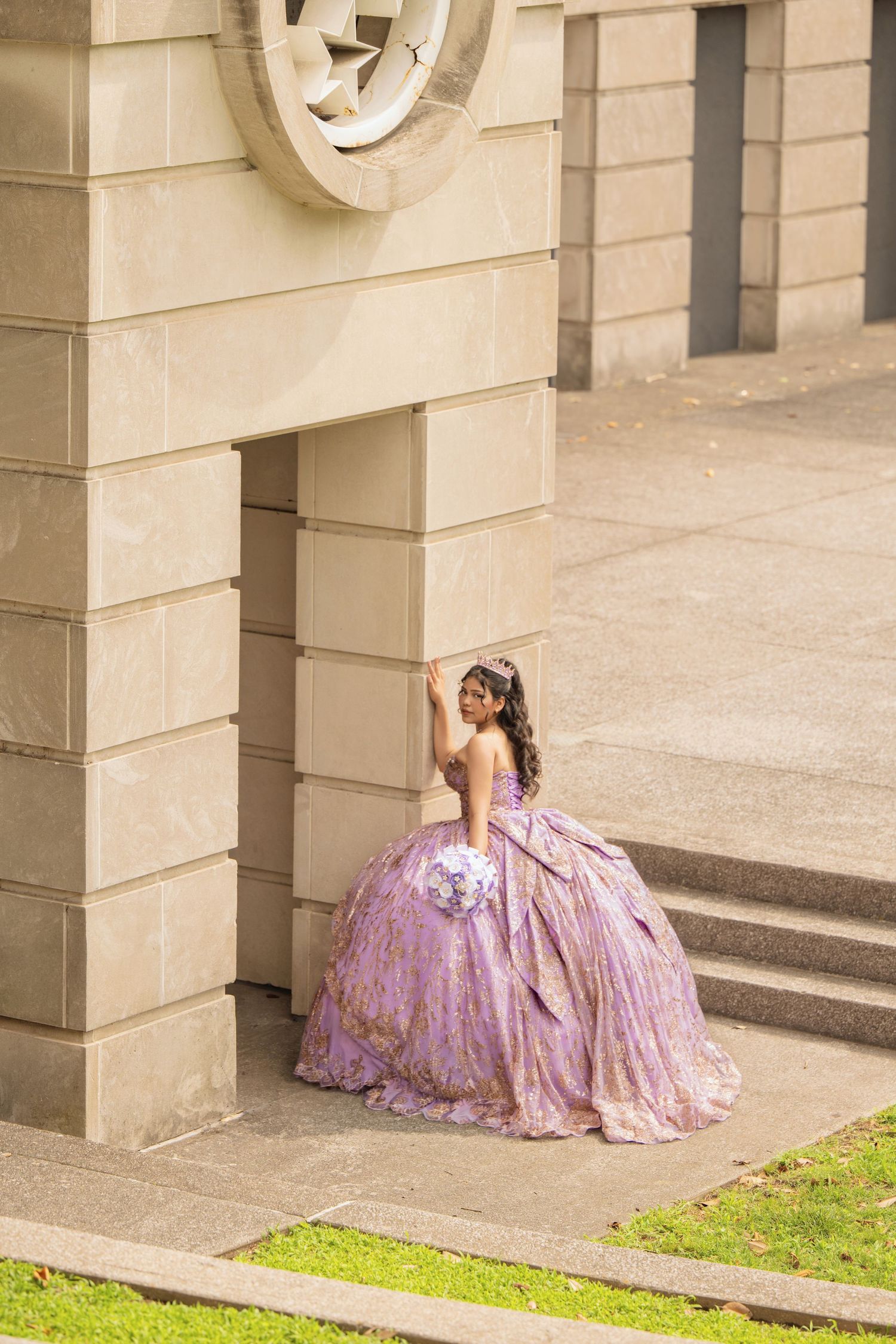 Young woman in lavish purple quinceañera ball gown poses gracefully beside ornate stone building columns.