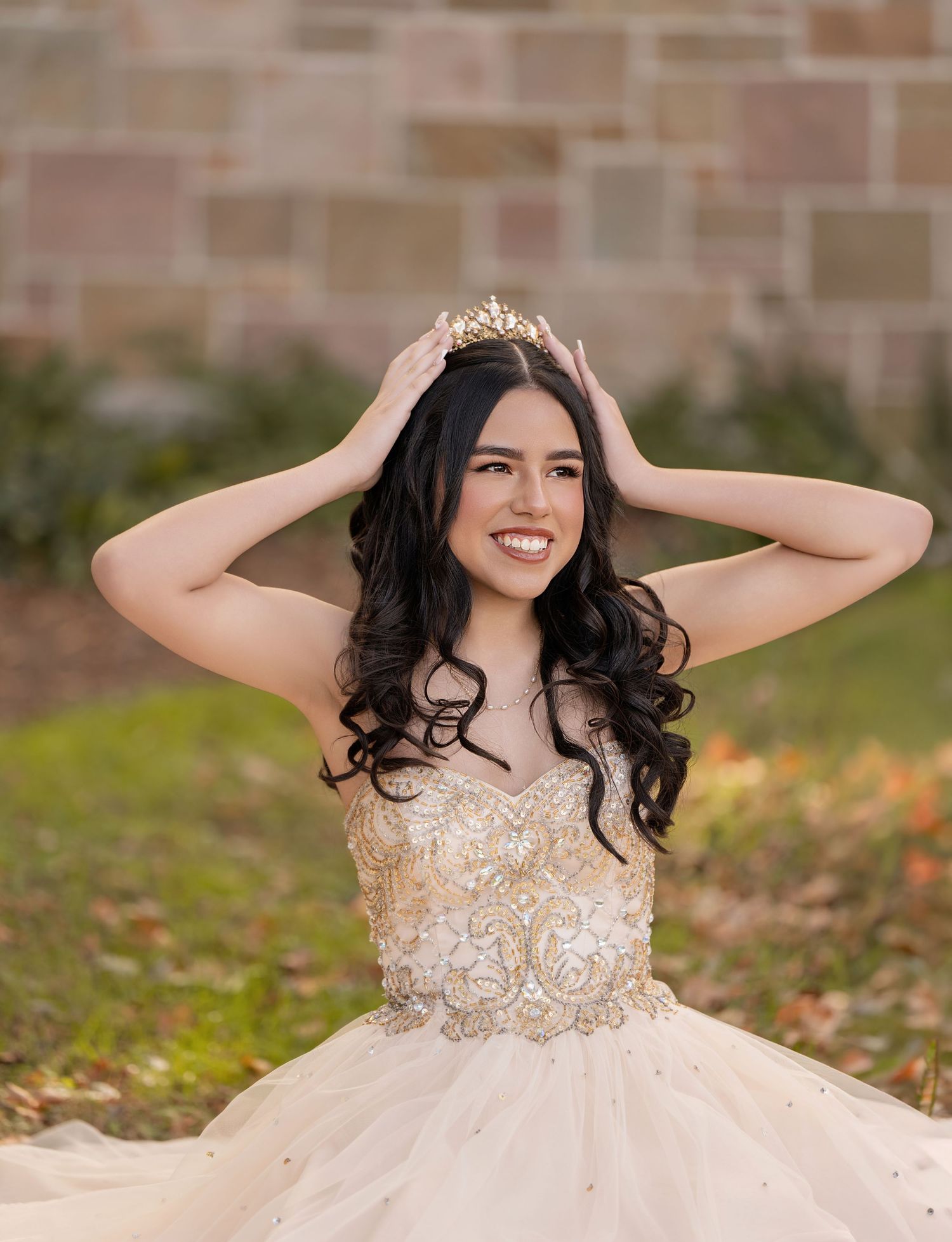 Young woman in gold beaded quinceañera gown adjusting tiara crown, smiling outdoors near stone wall.