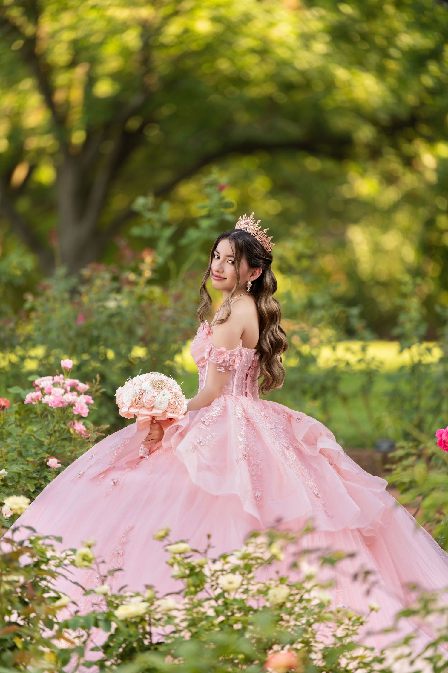 Young woman in pink ball gown with floral crown holds bouquet among blooming roses in lush garden.