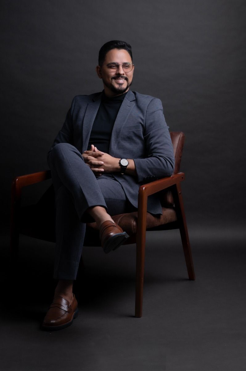Professional man in gray suit sitting confidently in a wooden chair, smiling, wearing a watch and brown loafers.