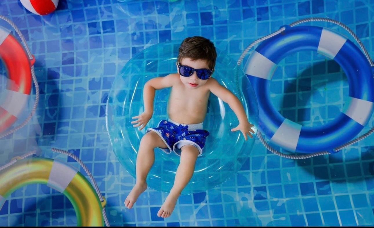 Young swimmer in blue swim trunks and sunglasses floating in a pool surrounded by colorful pool floats.