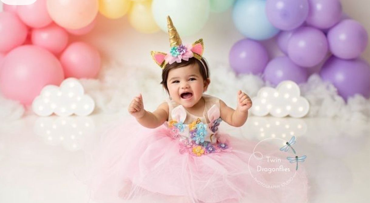 A baby in a pink tutu dress and unicorn headband sits against a pastel balloon backdrop during a birthday photoshoot.
