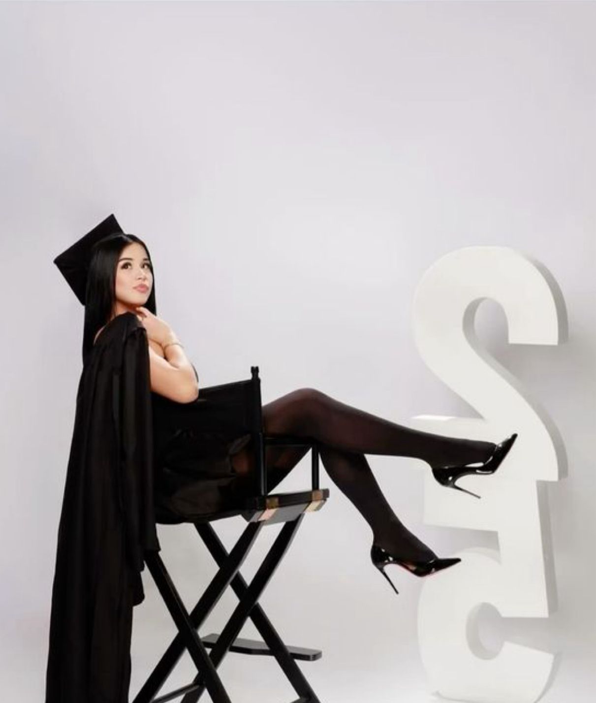 Graduate in black cap and gown sits on director's chair with crossed legs against white background with decorative letter S.