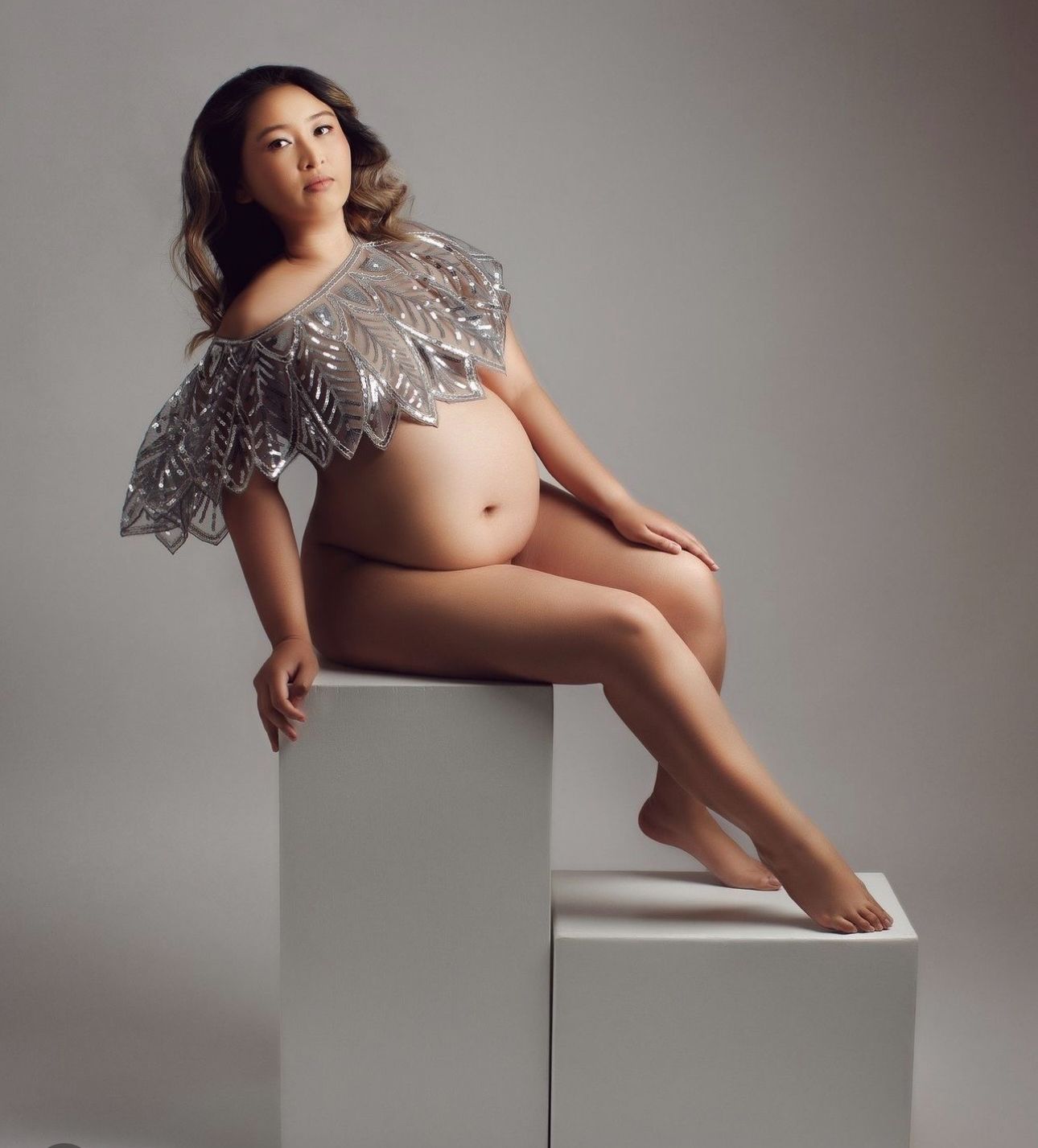 Pregnant woman posing elegantly on white cube wearing silver leaf cape in professional maternity studio portrait.
