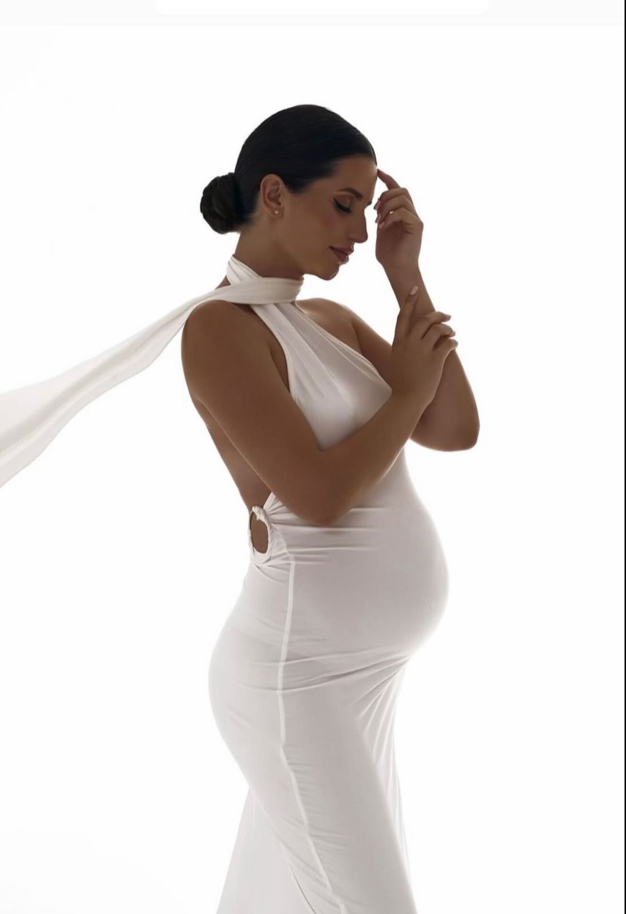 Pregnant woman in elegant white halter gown with flowing cape, posing gracefully against white background.
