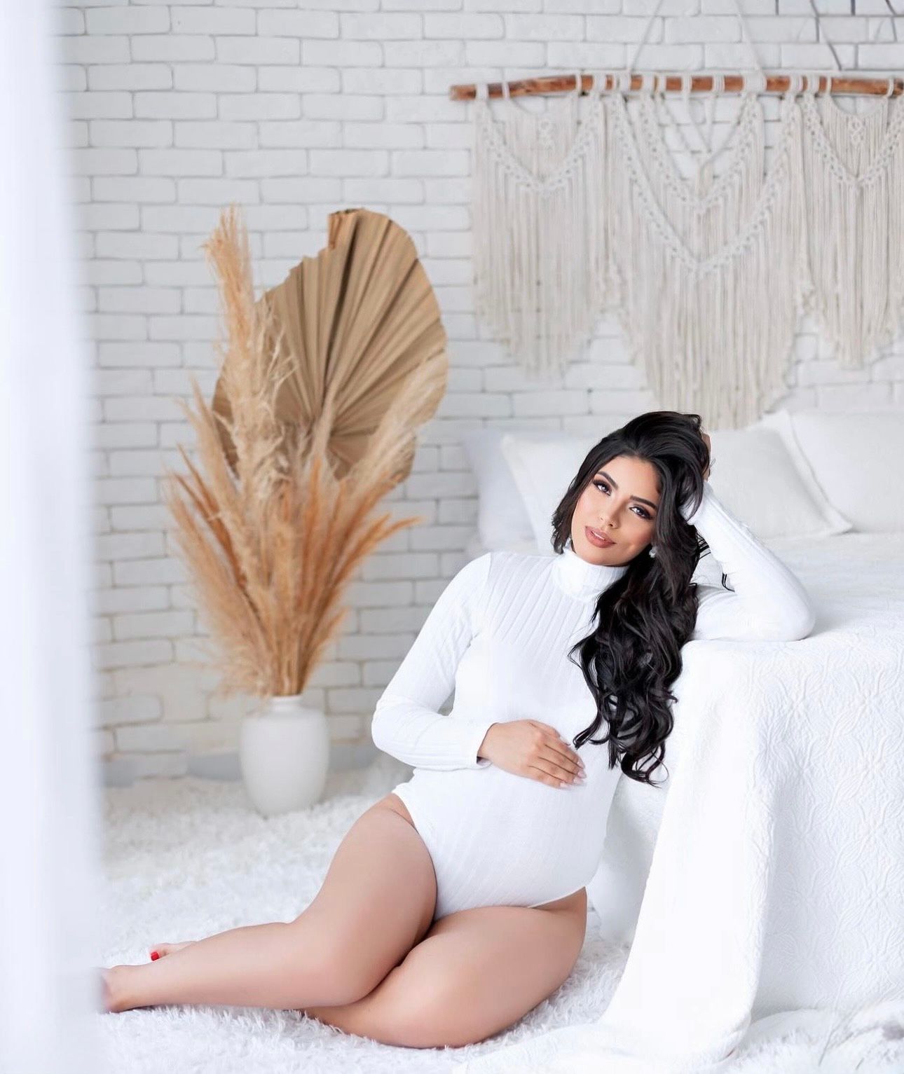 Pregnant woman in white bodysuit relaxing on floor beside bed in boho-styled bedroom.