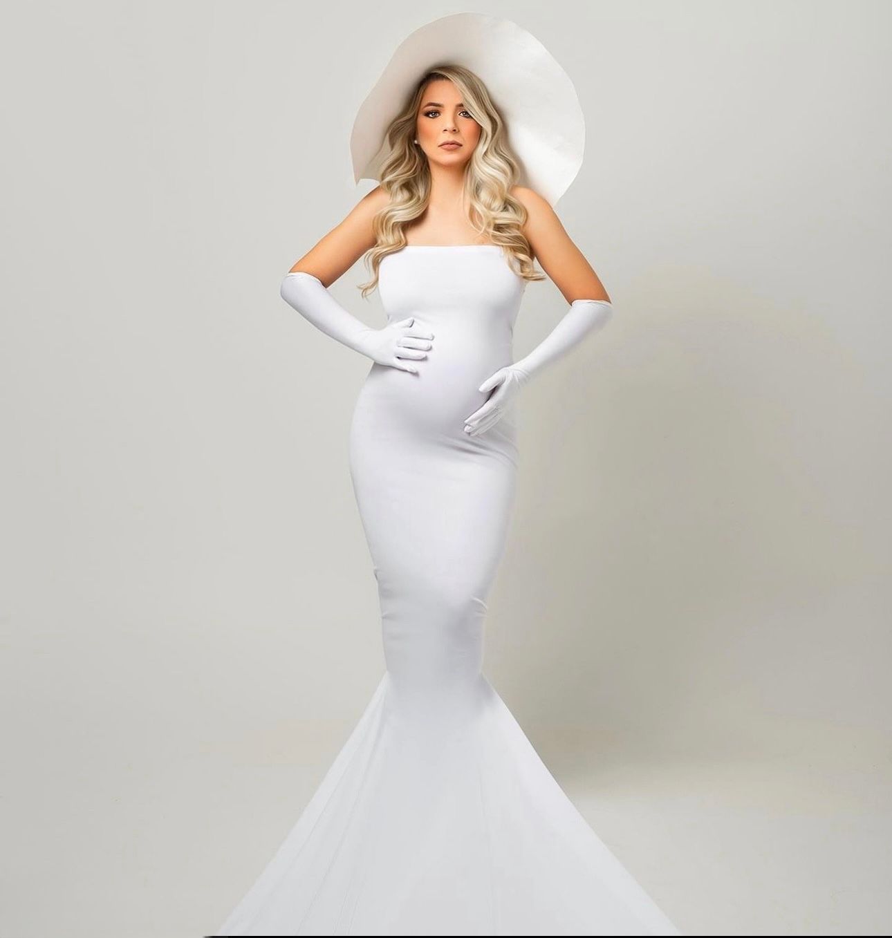 Pregnant woman in elegant white strapless mermaid gown with wide-brim hat and long gloves for maternity shoot.
