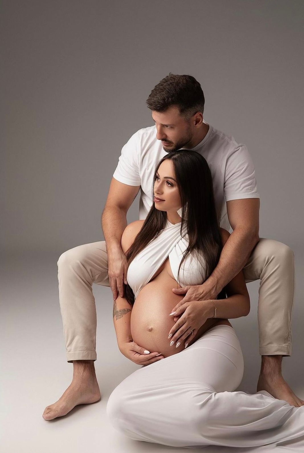 Couple maternity photo, pregnant woman in white dress cradled by partner on gray studio background.