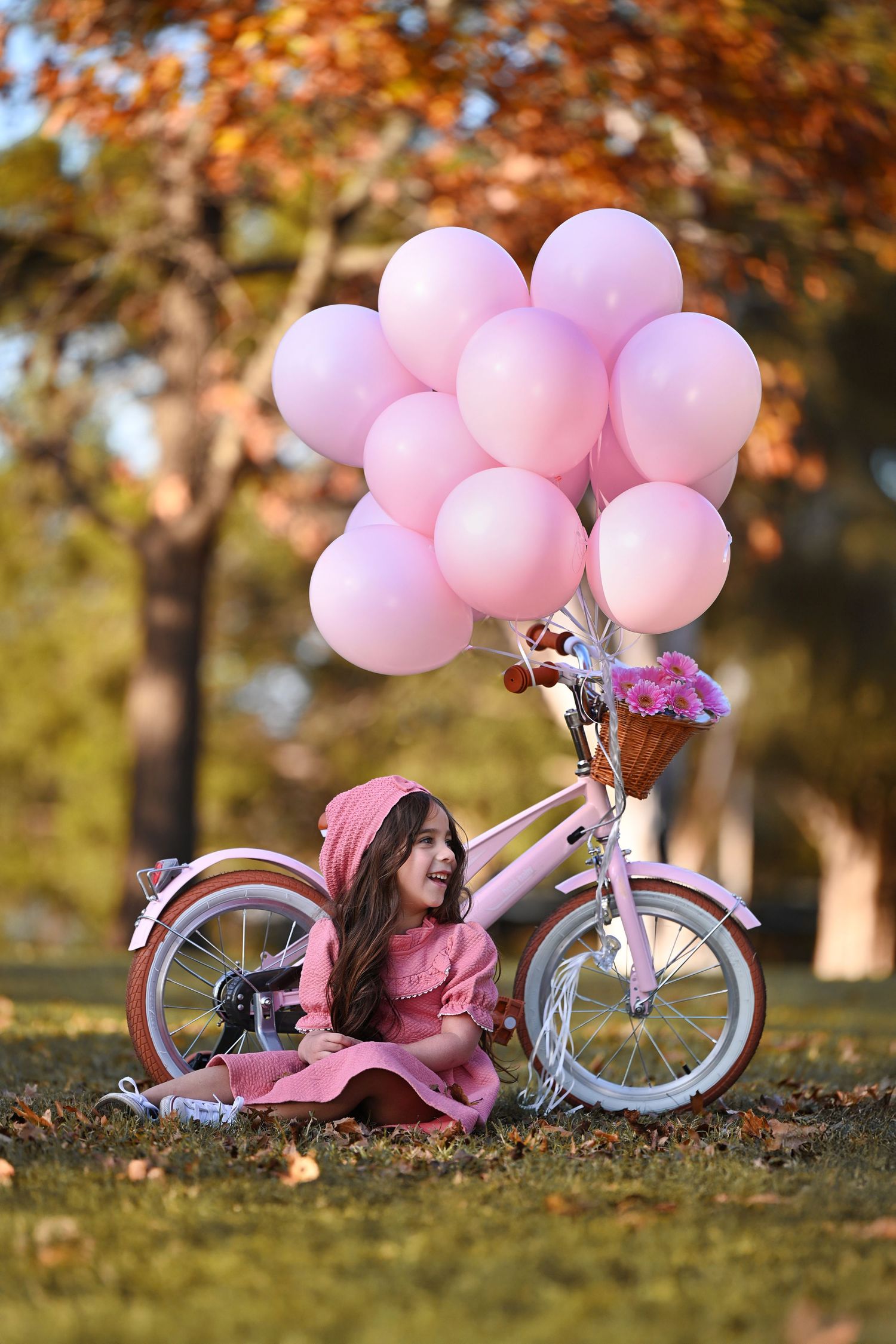 A little girl in a pink dress sits on grass beside a white bicycle adorned with pink balloons and flowers in a park.
