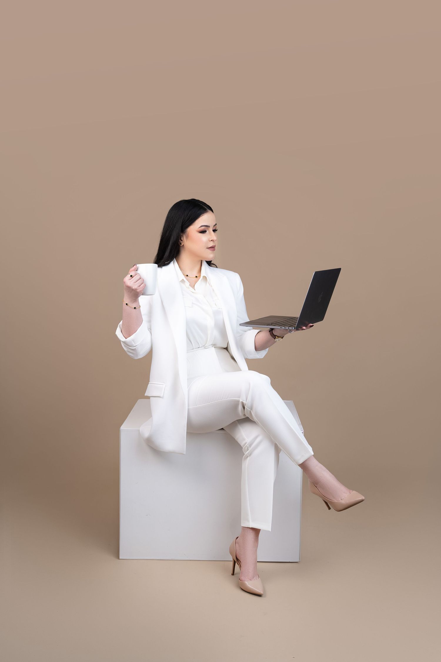 Professional woman in white suit sitting on cube, holding laptop and coffee cup against beige background.