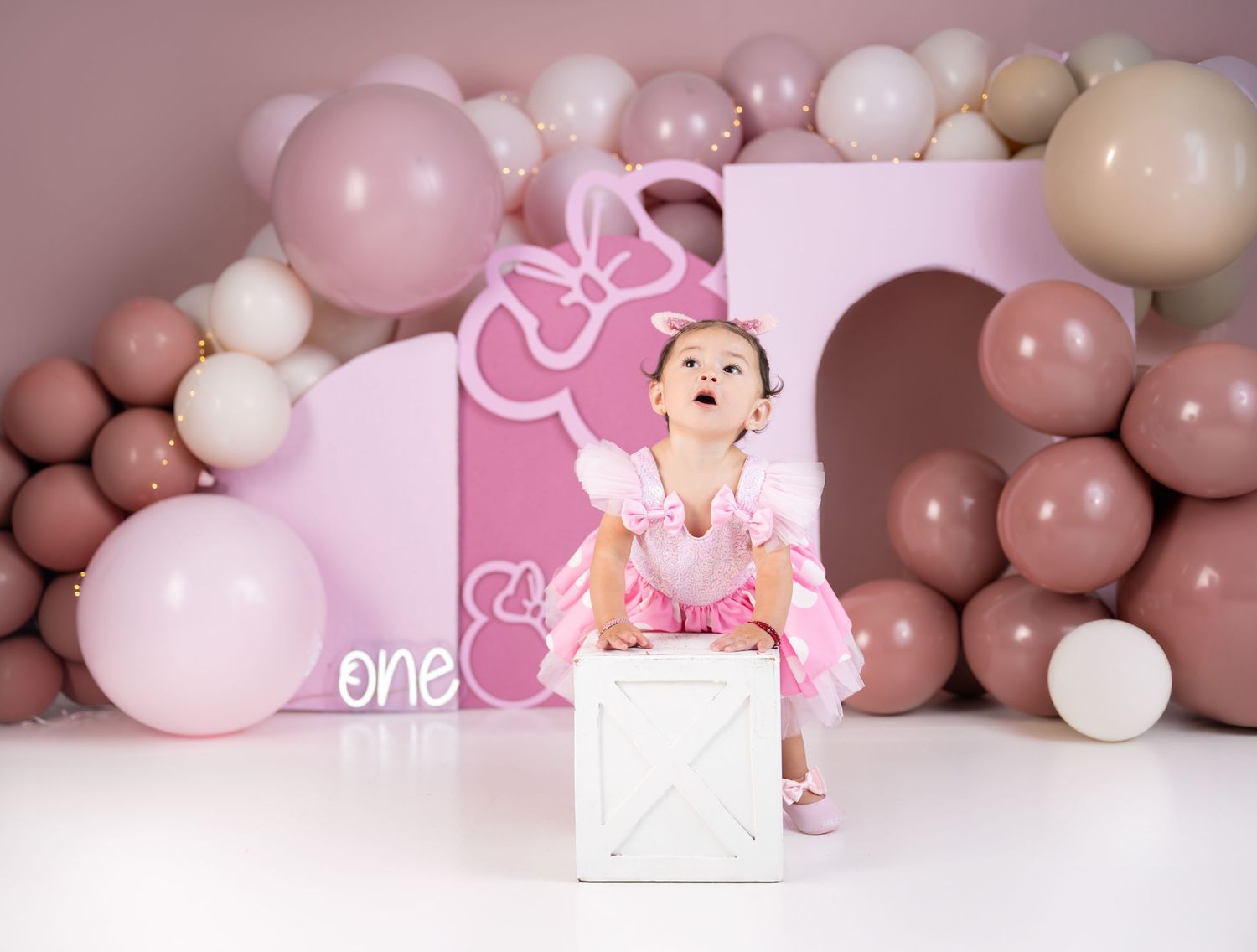 Baby girl in pink dress sits on white box at Minnie Mouse first birthday cake smash with balloon backdrop.