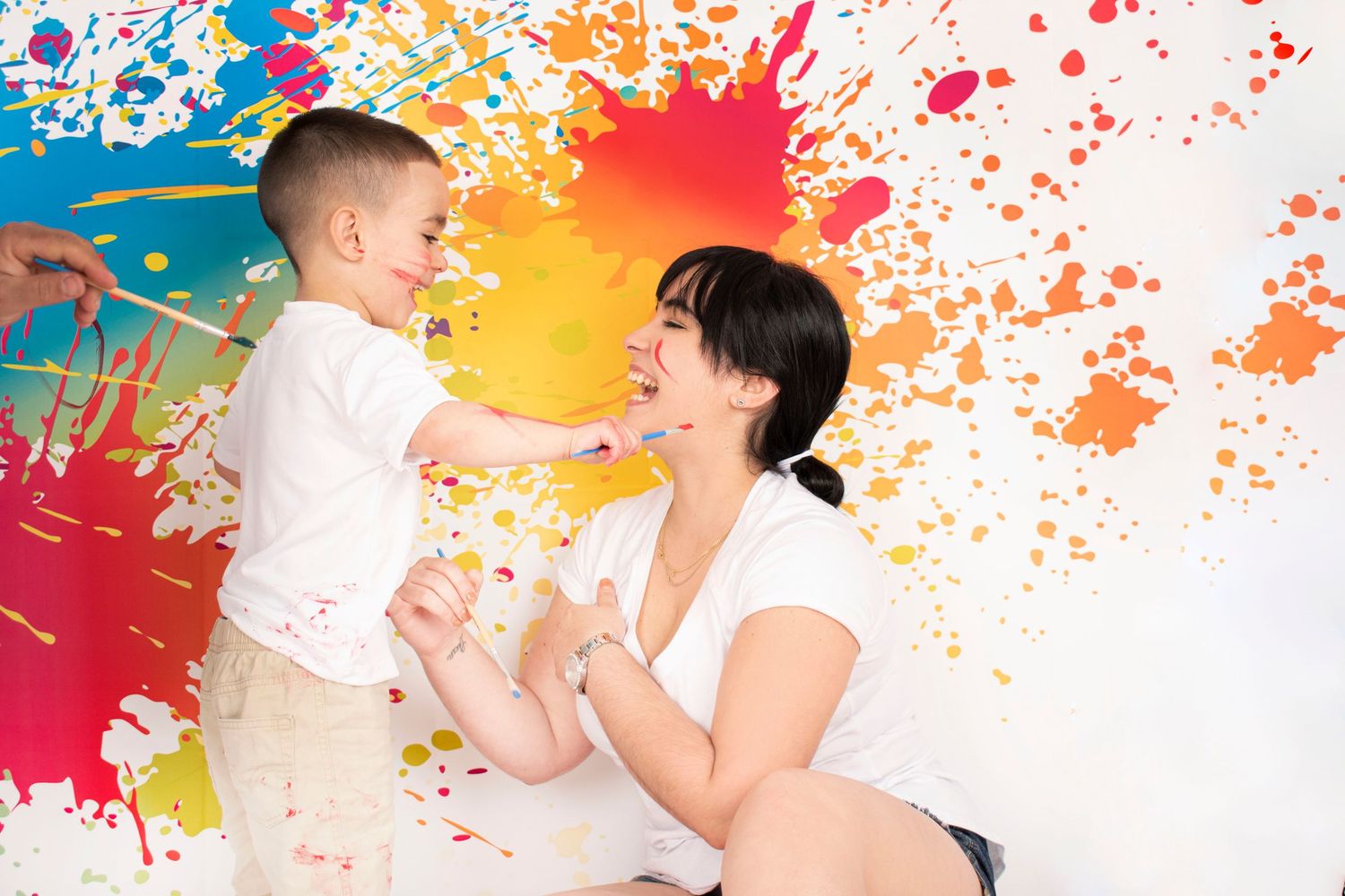 A joyful mother and young son play together against a colorful paint splatter wall background.