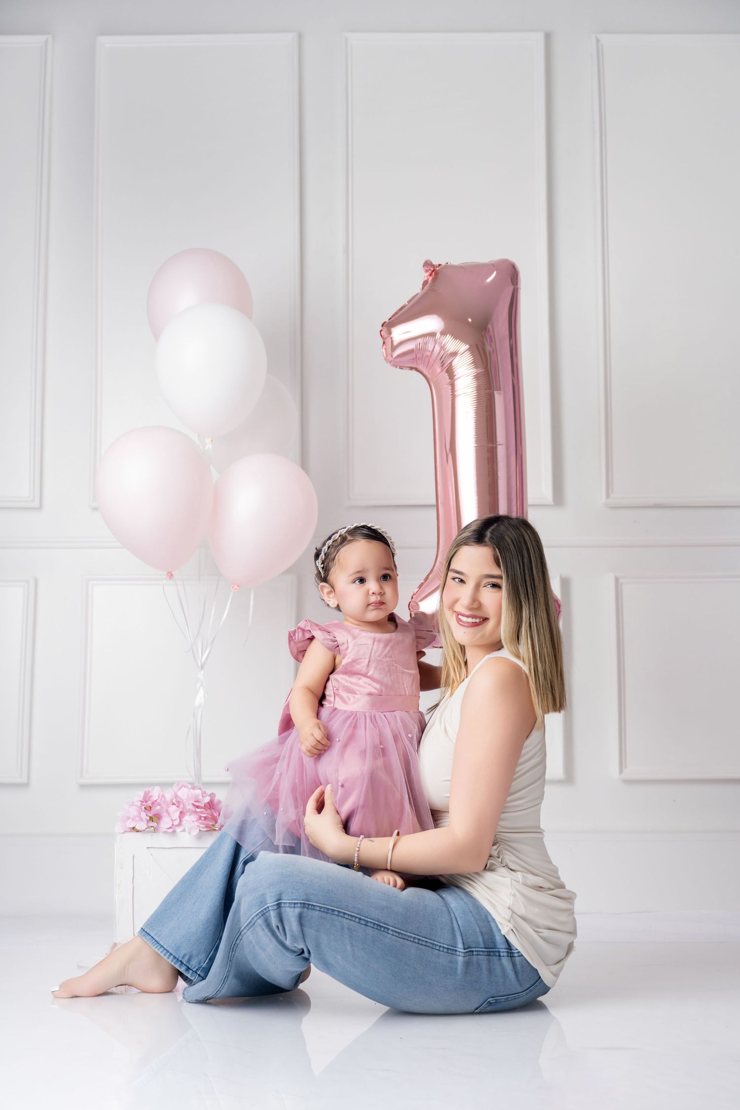 Mother and baby girl in pink dress celebrate first birthday with balloons and number one decoration.