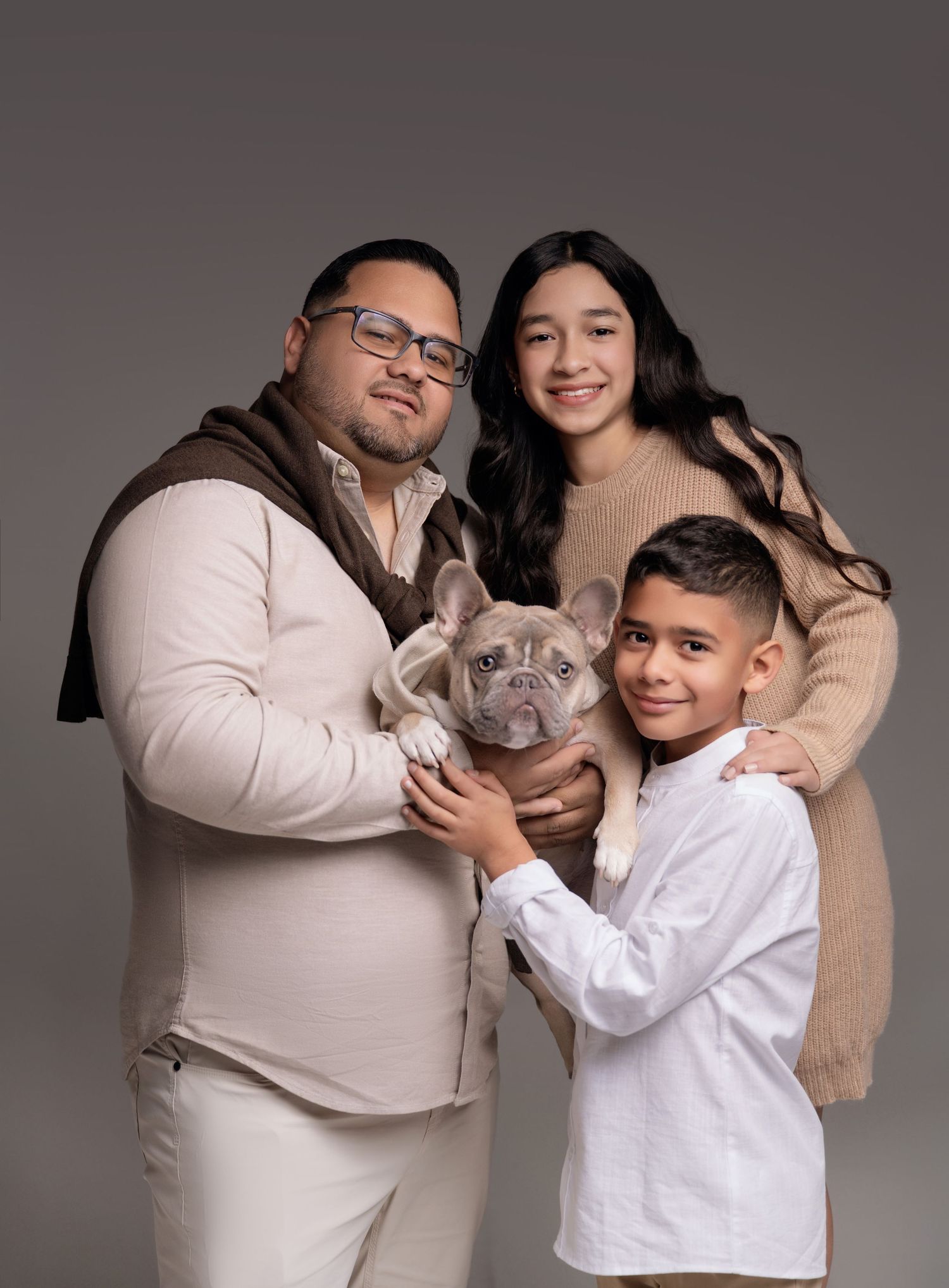 Family portrait of two adults and a boy holding a French Bulldog, dressed in neutral tones against a gray backdrop.