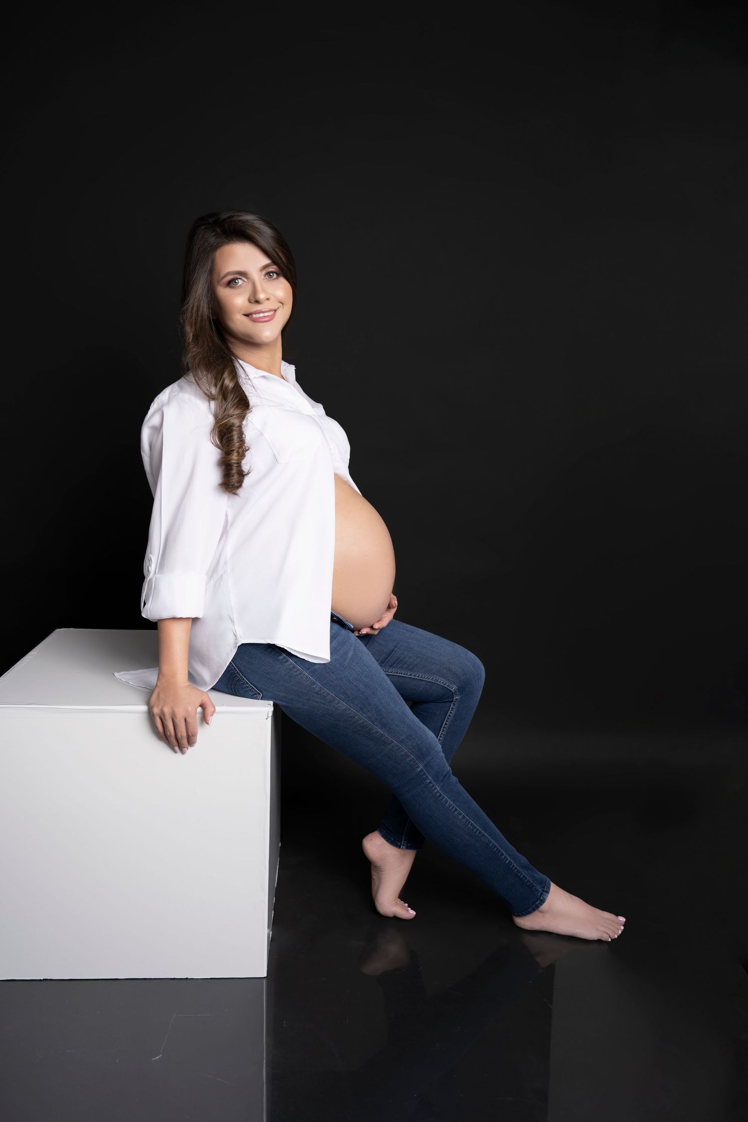 Pregnant woman in white shirt and jeans sitting on white cube, smiling, against black background.