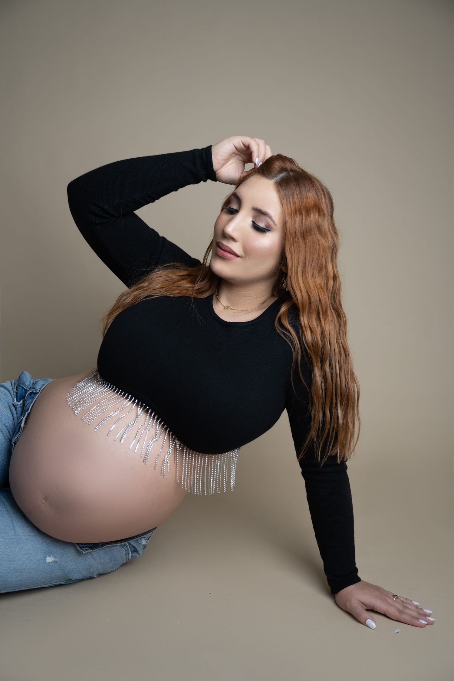 Pregnant woman with long red hair in black top and jeans, posing on floor with baby bump exposed.
