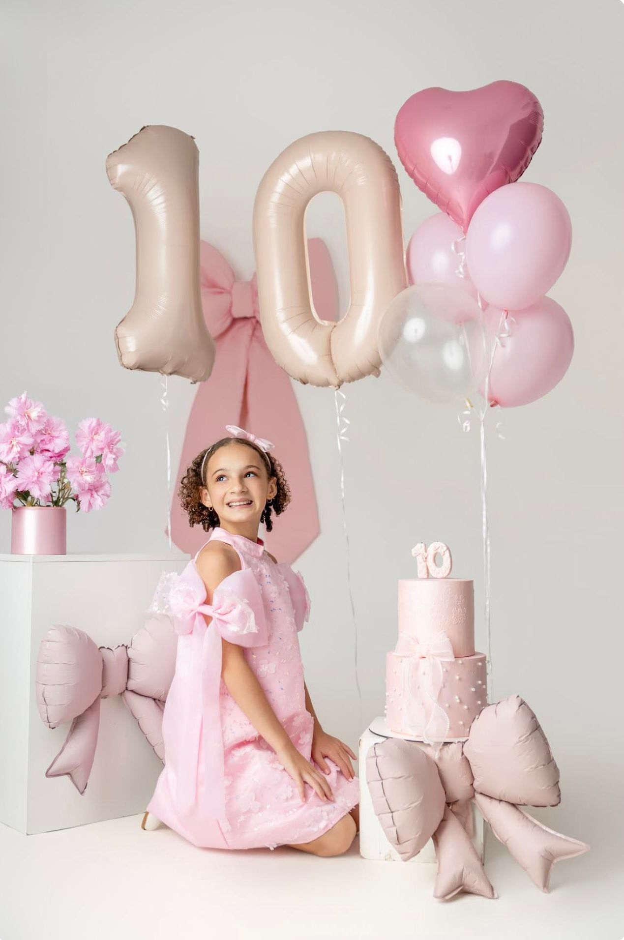 A smiling girl in a pink dress celebrates her 10th birthday with balloons, a tiered cake, and pink decorations.