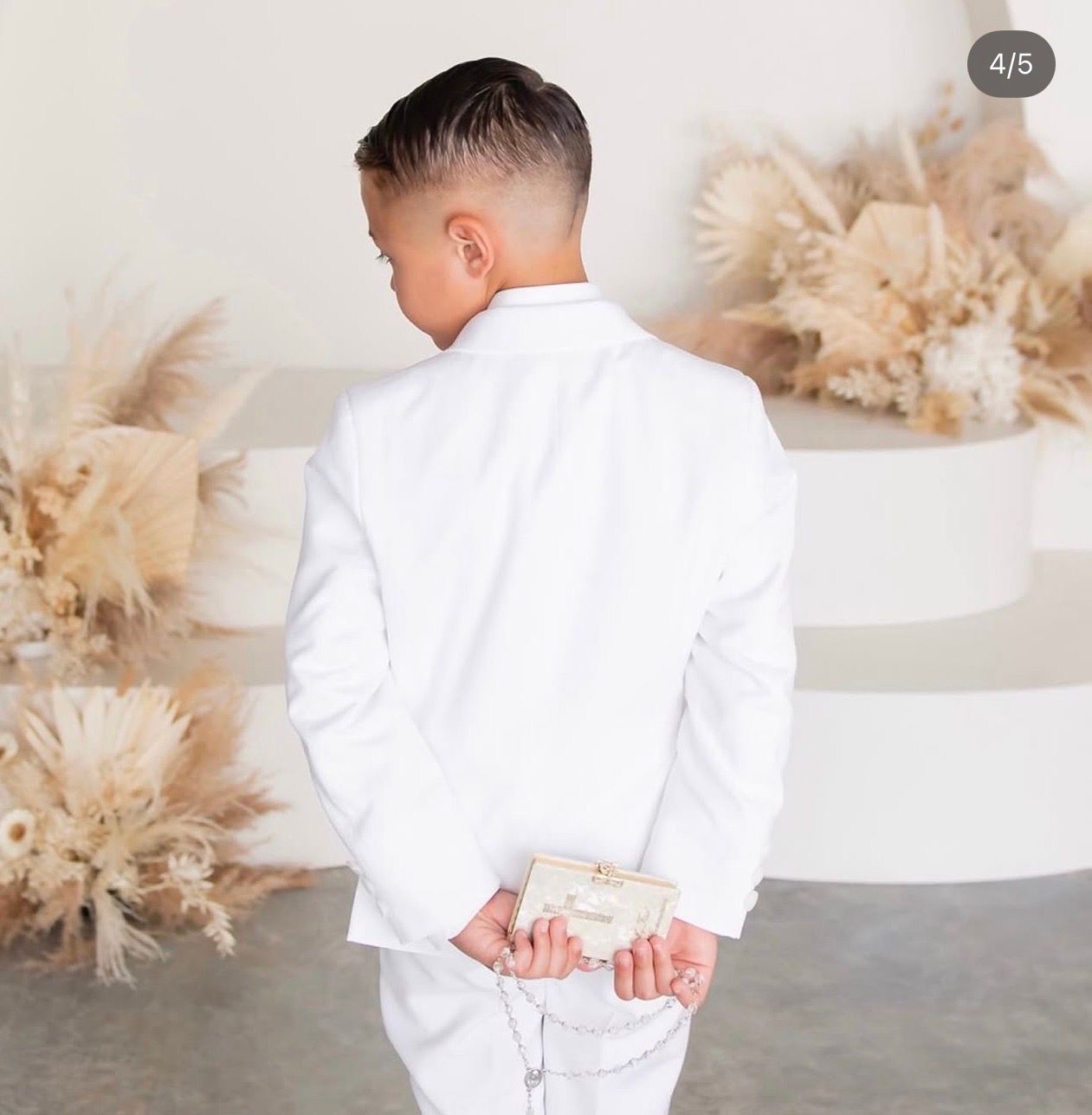 Young boy in white suit stands with hands behind back near floral arrangement and cake.