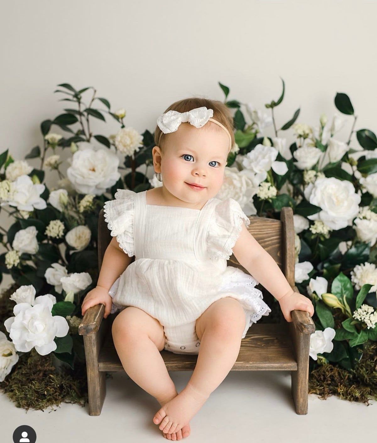 Adorable baby girl in white romper and bow headband sitting on wooden chair surrounded by white flowers.