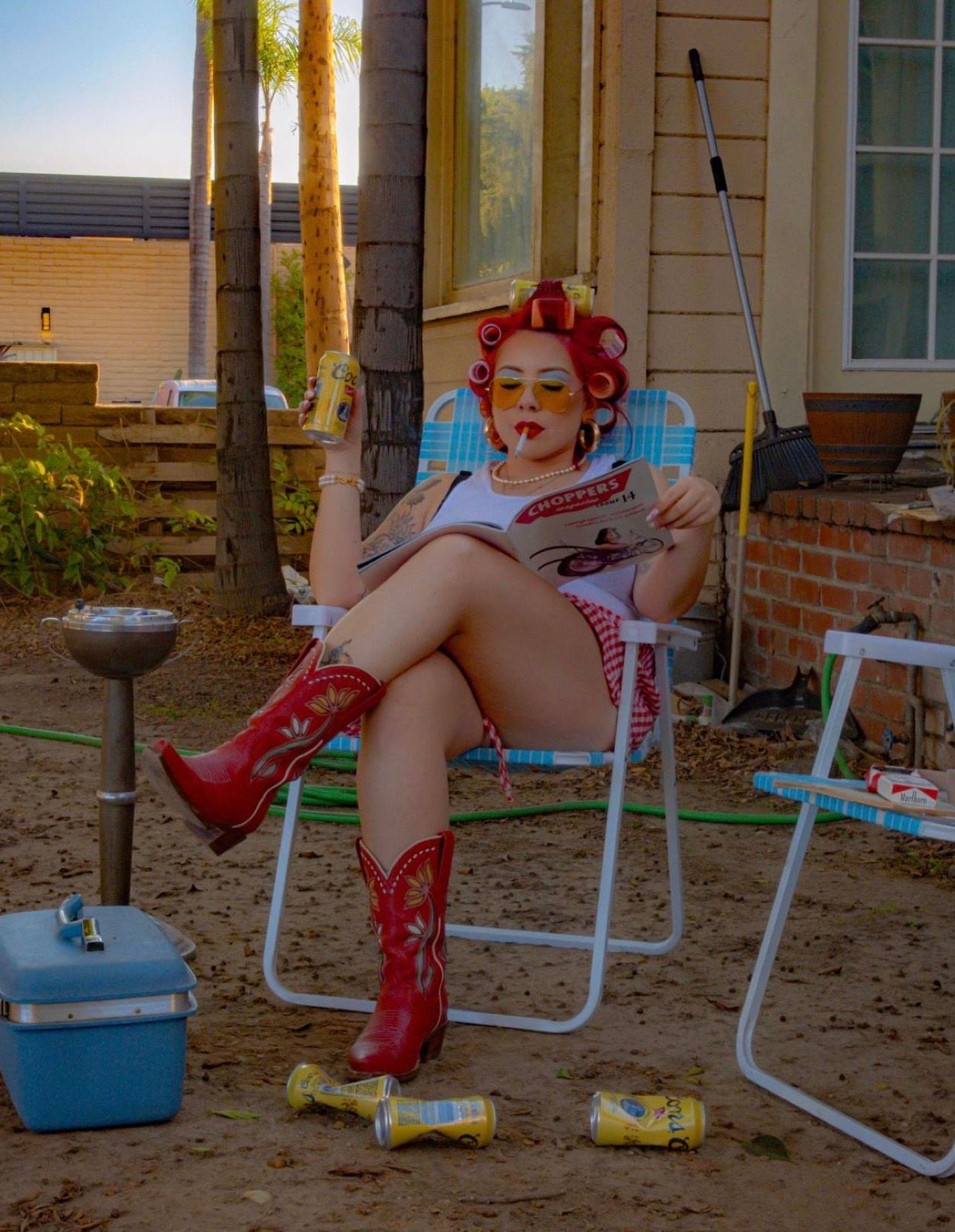Woman in red curlers and cowboy boots relaxing in lawn chair reading magazine outside house.