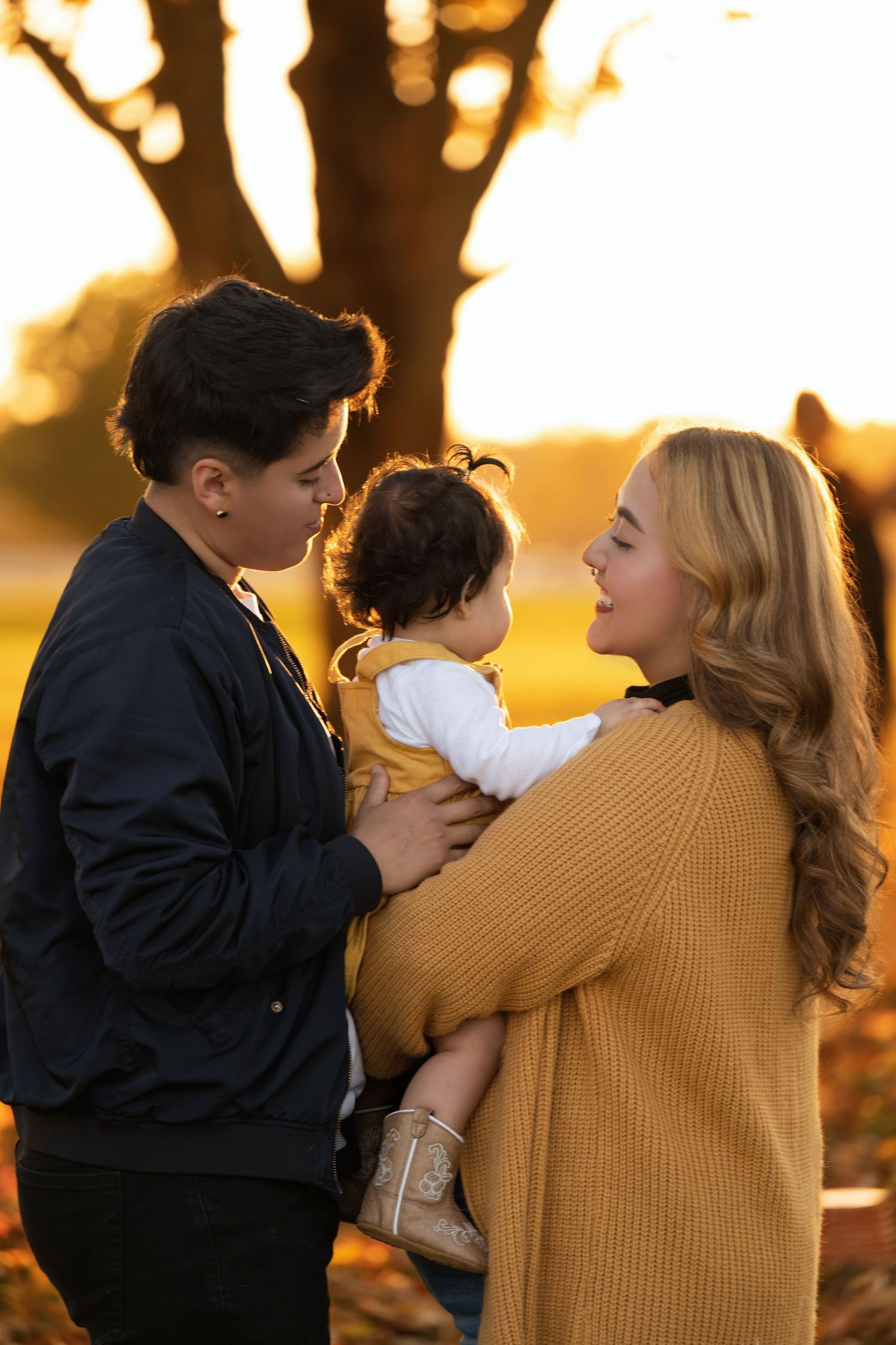 Two parents sharing a tender moment with their baby at golden hour sunset in an autumn outdoor setting.