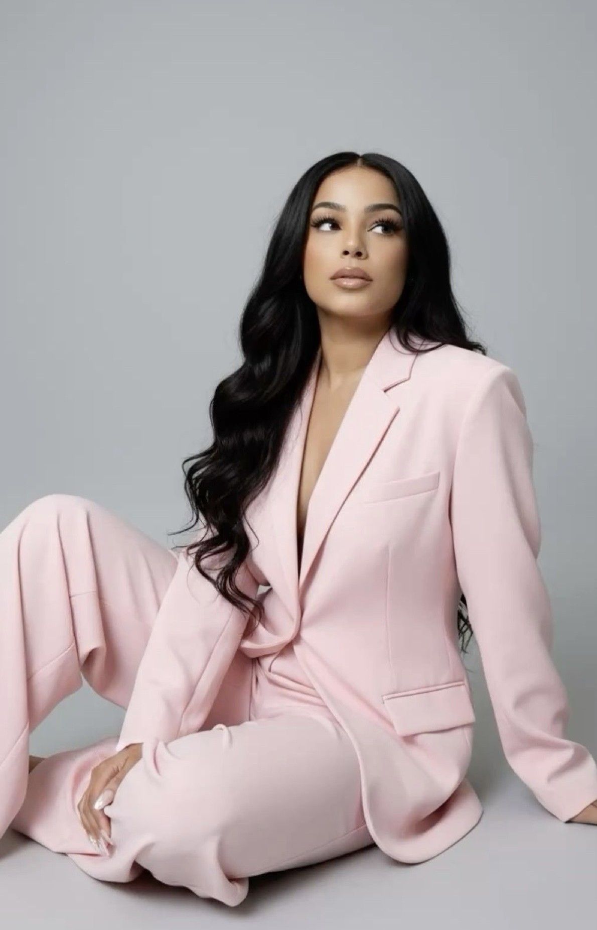 Woman in pink blazer suit with long black hair sitting gracefully against a gray background.