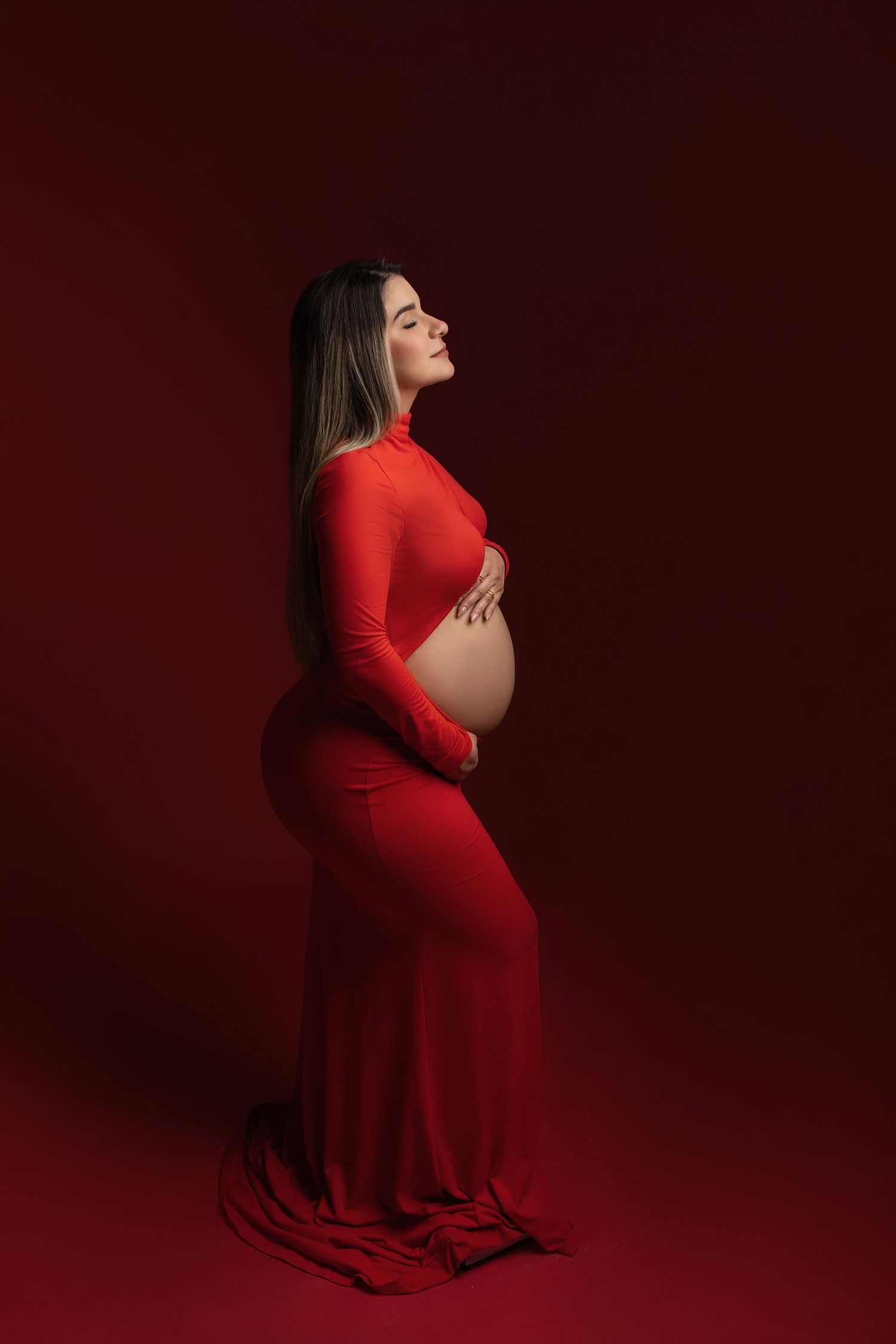 Artistic maternity photo series featuring a woman in a flowing red dress against a dark burgundy backdrop.