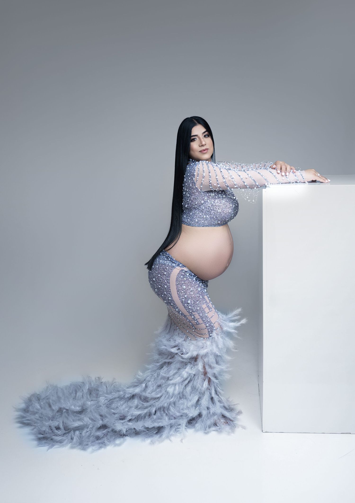 A maternity photoshoot featuring a glamorous grey tulle gown with feathers against a light grey studio backdrop.