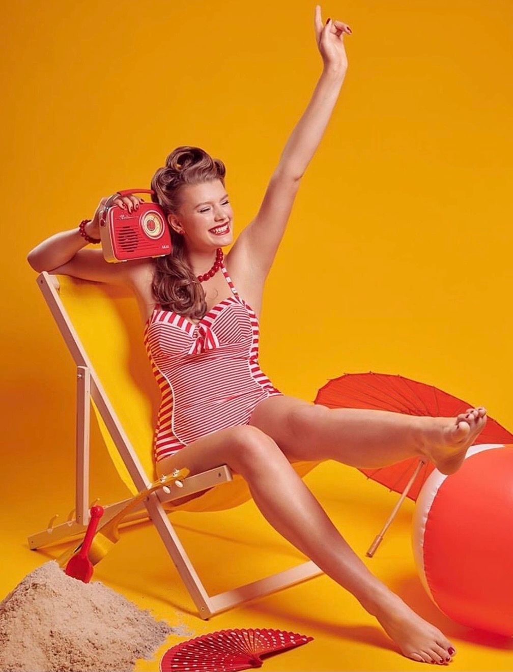 Retro pin-up woman in red striped swimsuit sitting in beach chair holding vintage radio on yellow background.