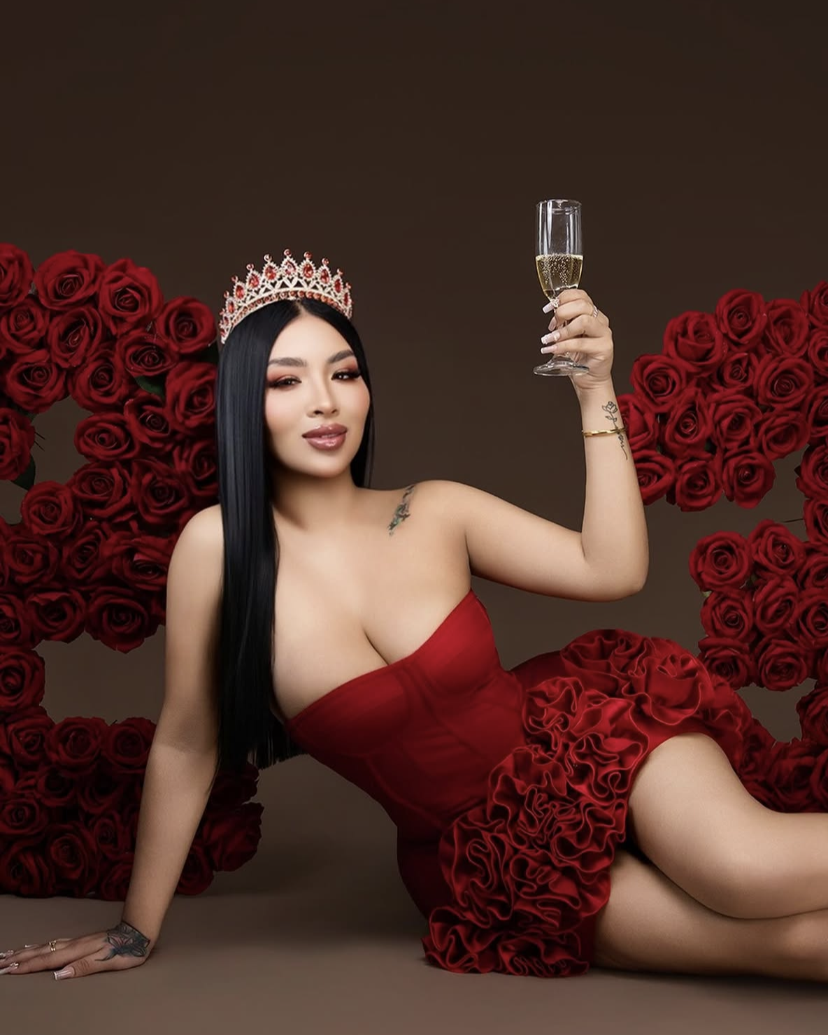 Woman in red dress and crown holds champagne glass, posed among red roses in elegant Valentine's Day themed photo.