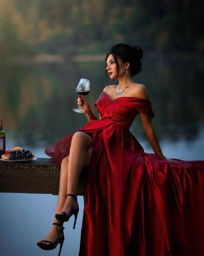 Elegant woman in red off-shoulder gown holding wine glass, seated by a serene lake at dusk.