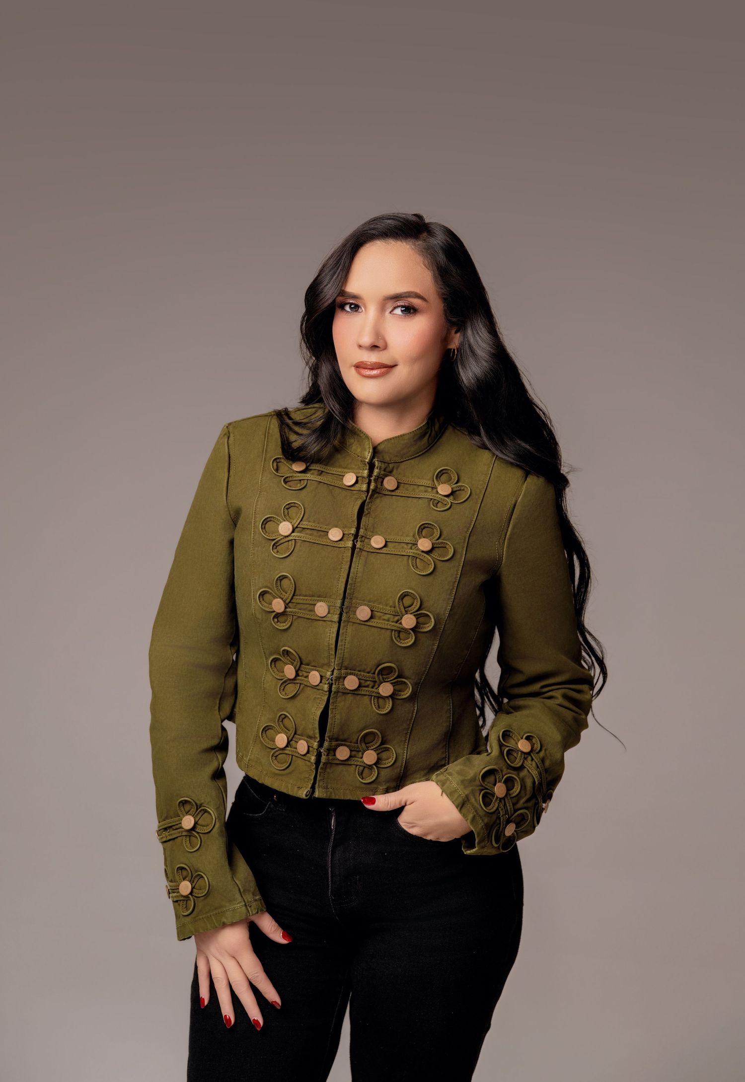 Woman wearing olive green military-style jacket with floral embellishments and gold buttons, paired with black jeans.