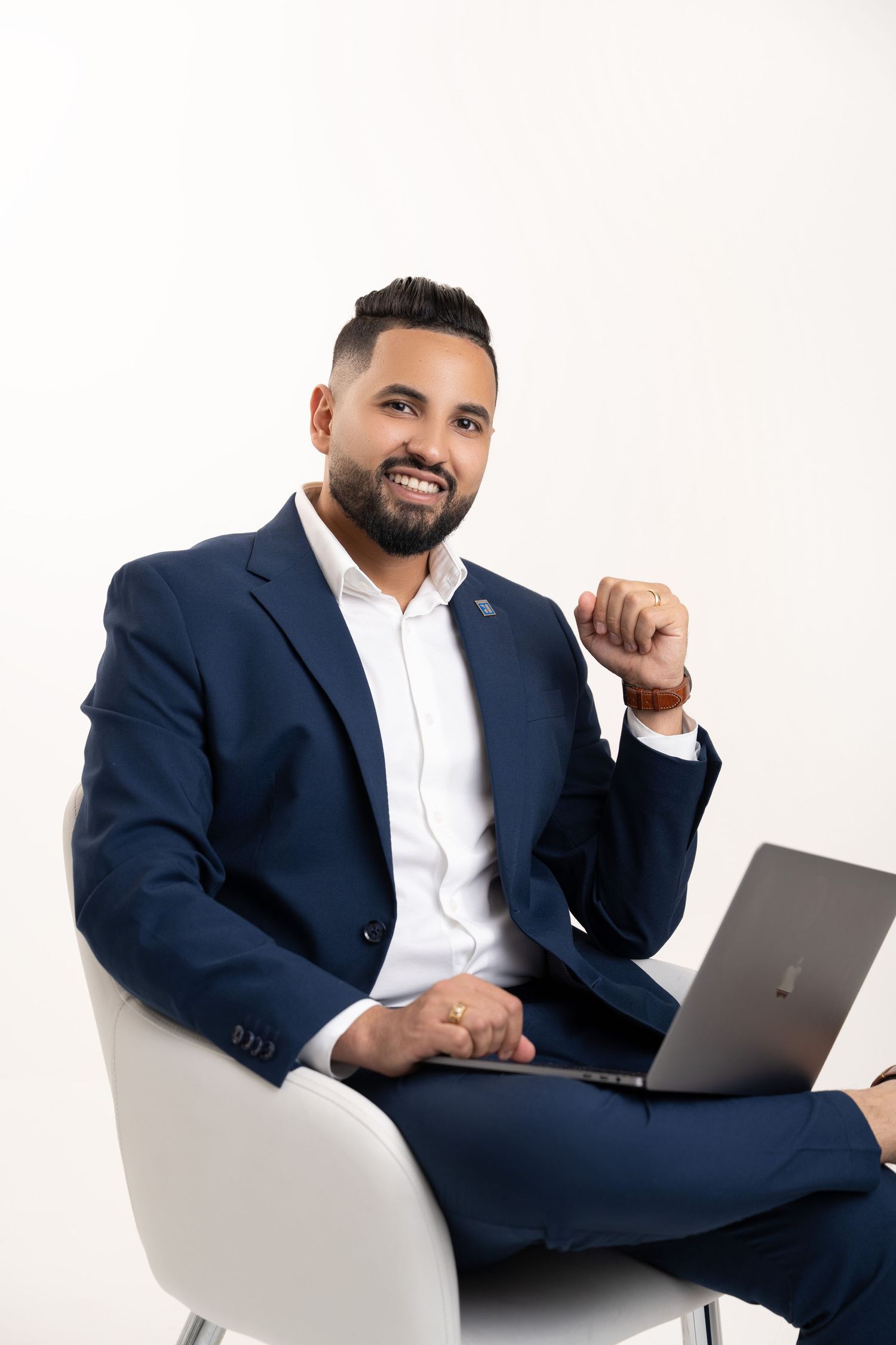 Smiling businessman in navy suit sitting in white chair with laptop, professional portrait on white background.