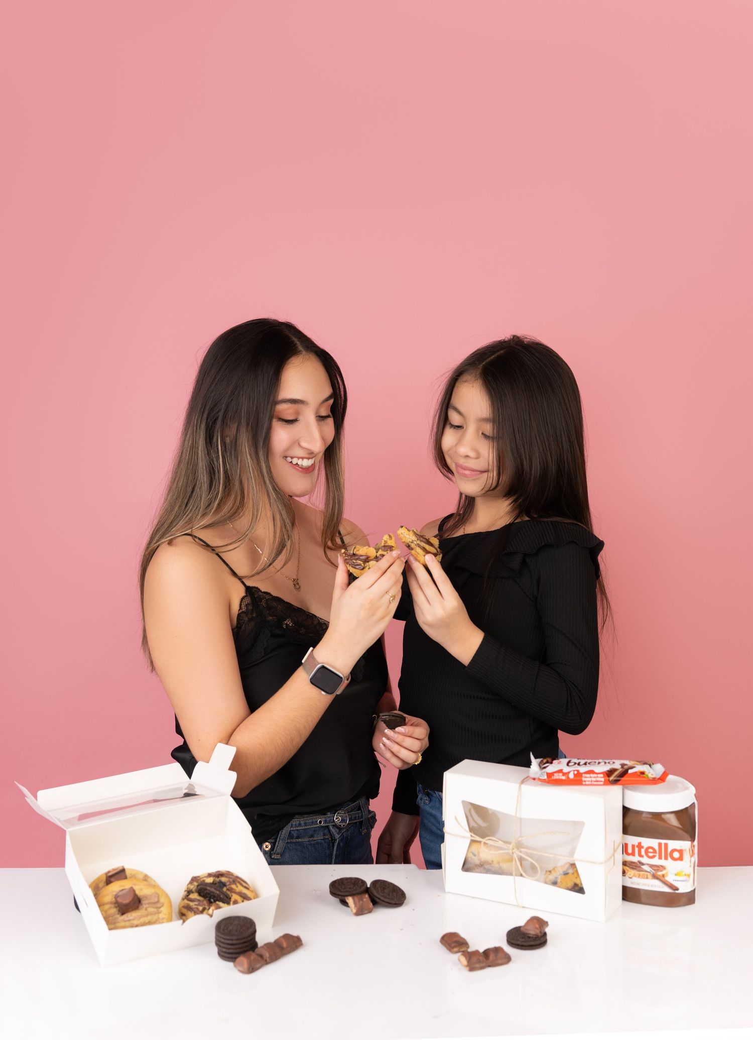 Woman and girl sharing a Nutella pastry treat at a white table with chocolate desserts and Nutella jar on pink background.