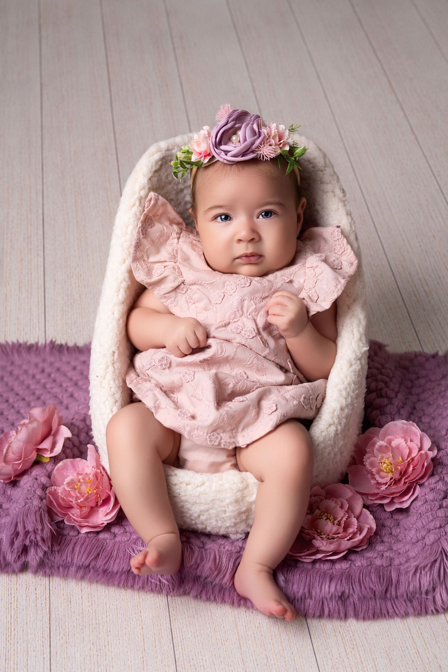 Baby girl in pink lace dress and floral crown sits in white chair on purple rug with pink flowers.
