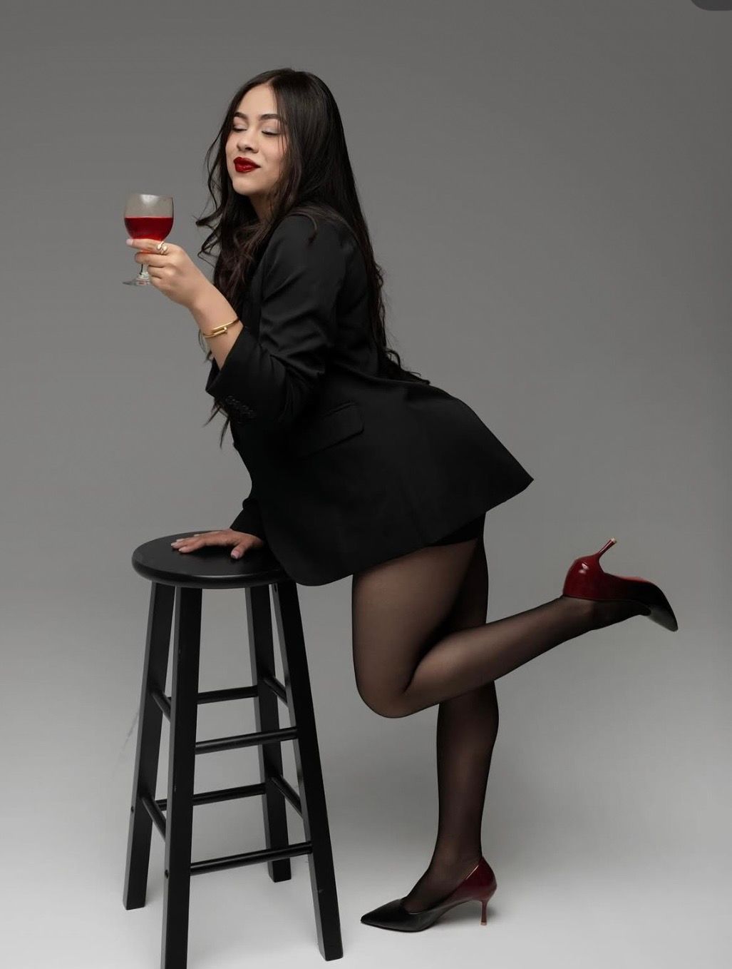 Woman in a black blazer dress, sheer tights and red heels holds a glass of red wine while leaning on a black stool in a studio.