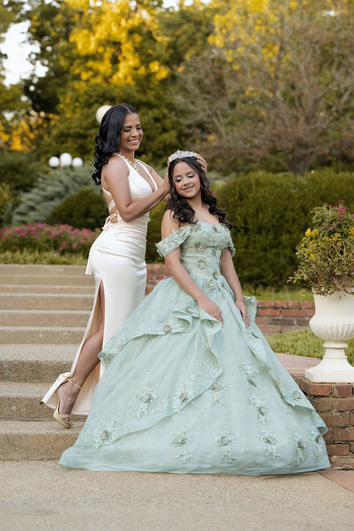 Two women pose outdoors, one in a white gown and one in a mint green quinceañera ball gown with floral details and tiara.