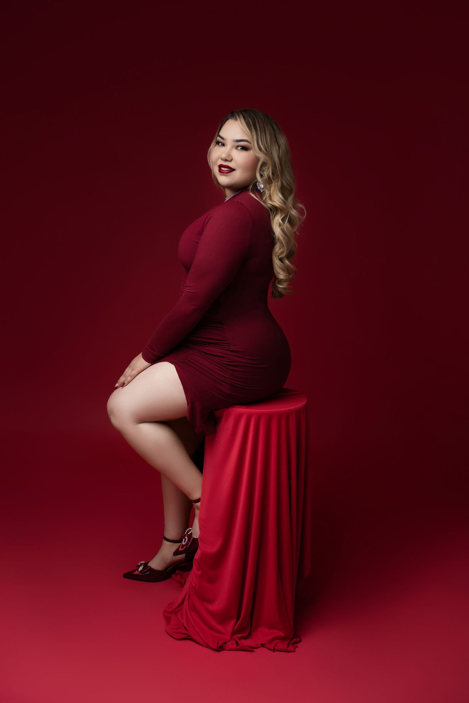 Woman with long wavy blonde hair in a burgundy long-sleeve dress sits on a draped red stool against a deep red studio backdrop.