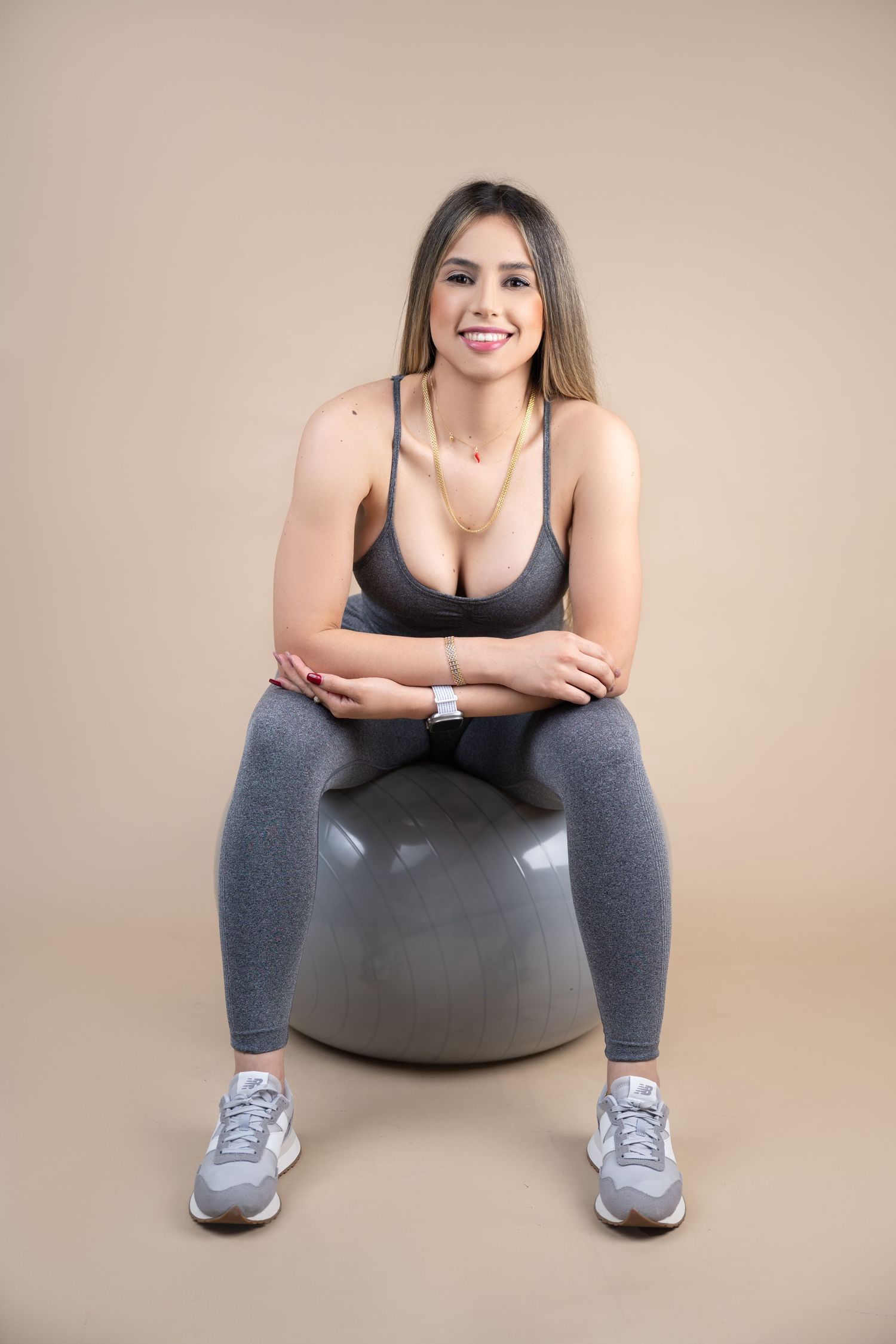 Smiling woman in gray athletic sports bra and leggings sits on a gray stability ball against a tan studio background.