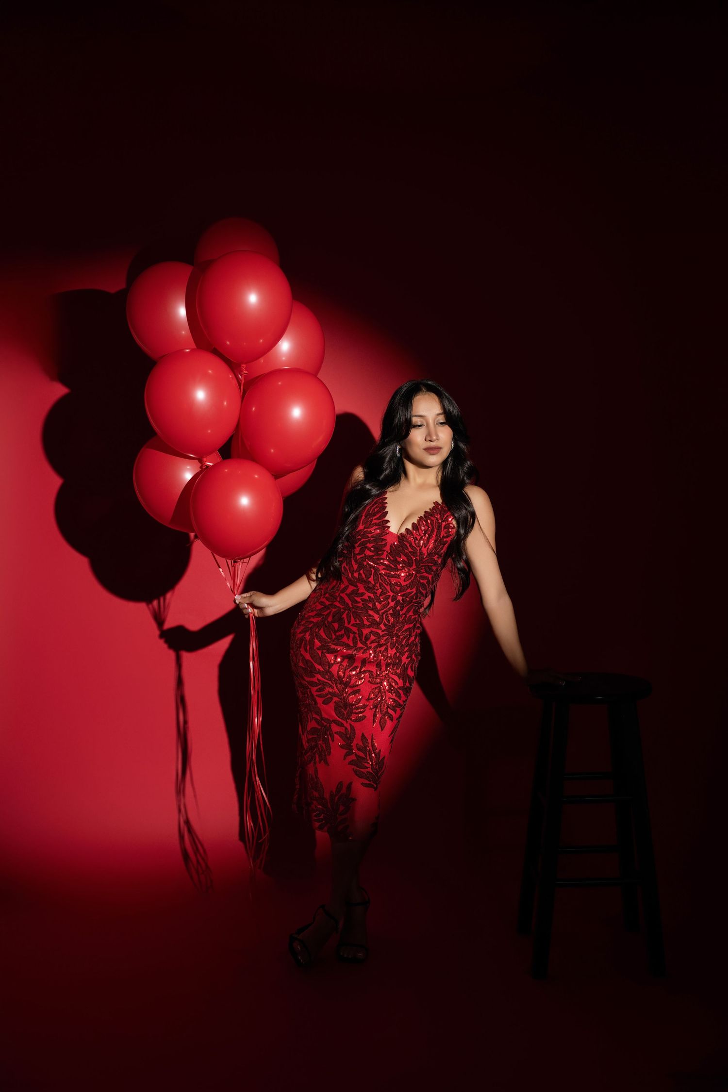 Woman in a sparkling red sequin dress holds a bunch of red balloons beside a black stool against a dramatic red spotlit backdrop.