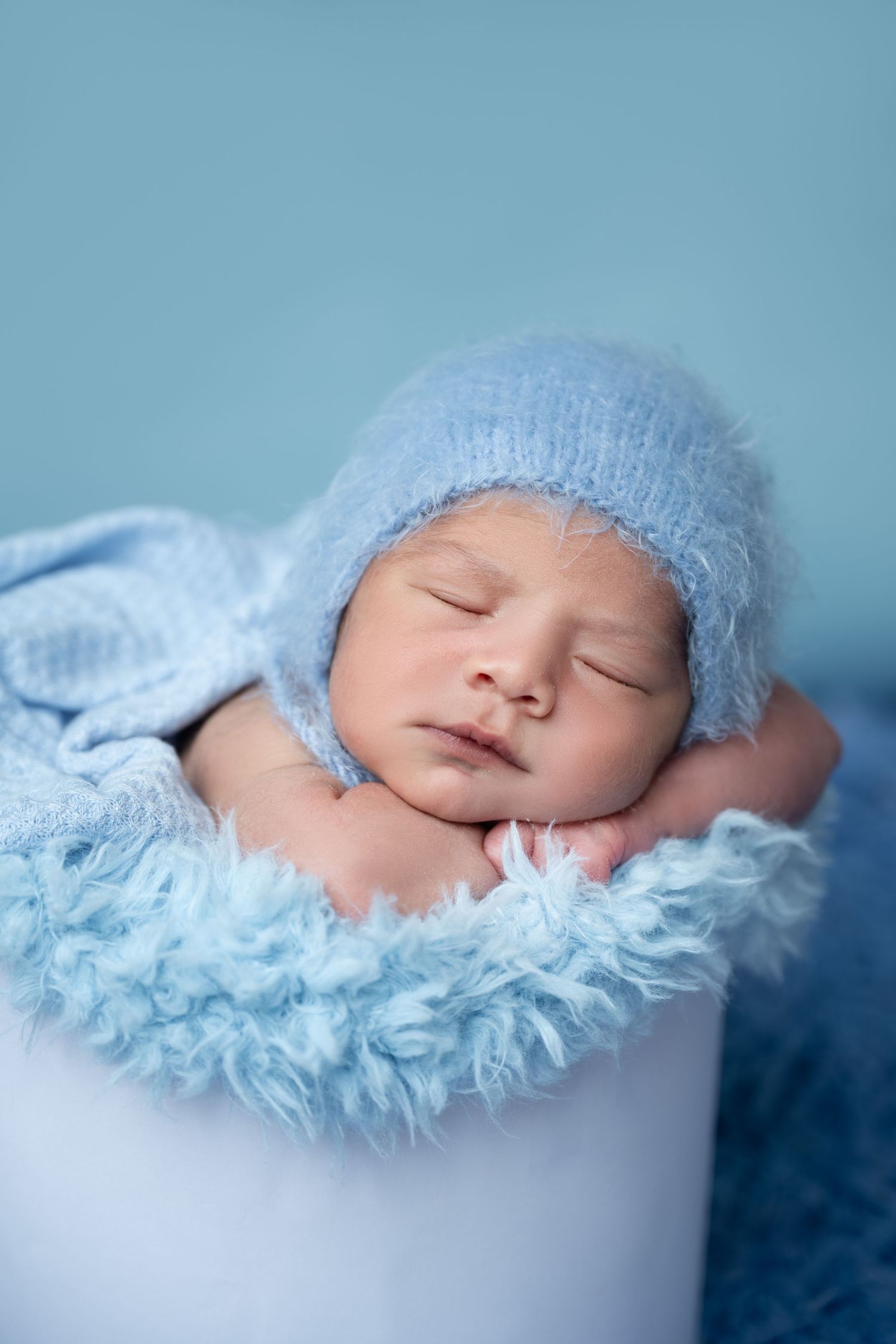 Sleeping newborn in blue knit hat resting in white container on blue textured backdrop.