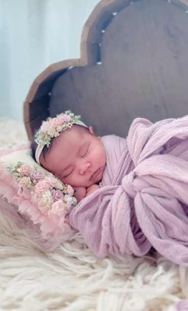A sleeping newborn baby wrapped in a lavender swaddle with small white flowers in their hair rests on soft white fur.