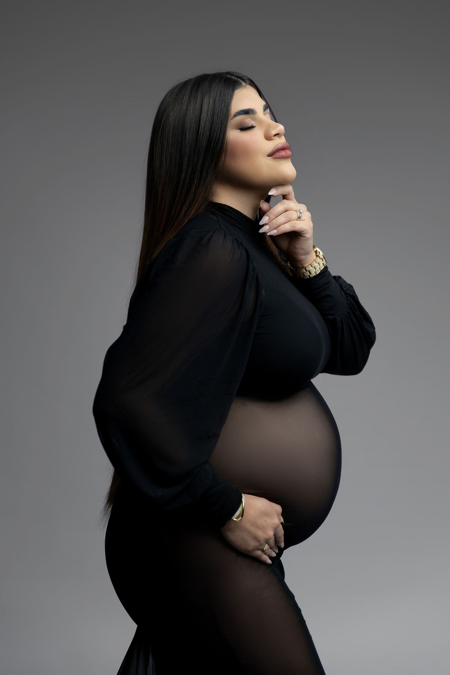 Elegant maternity portrait featuring a black sheer dress against a gray background in a dramatic side pose.