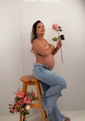 Maternity photo of expectant mother in jeans sitting on wooden stool holding pink rose against white backdrop.