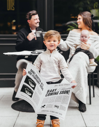Young child stands holding a newspaper with 'Baby Coming Soon!' headline while family relaxes at outdoor cafe.