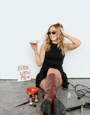 A person in a black dress and brown boots poses with a cocktail glass against a white wall.