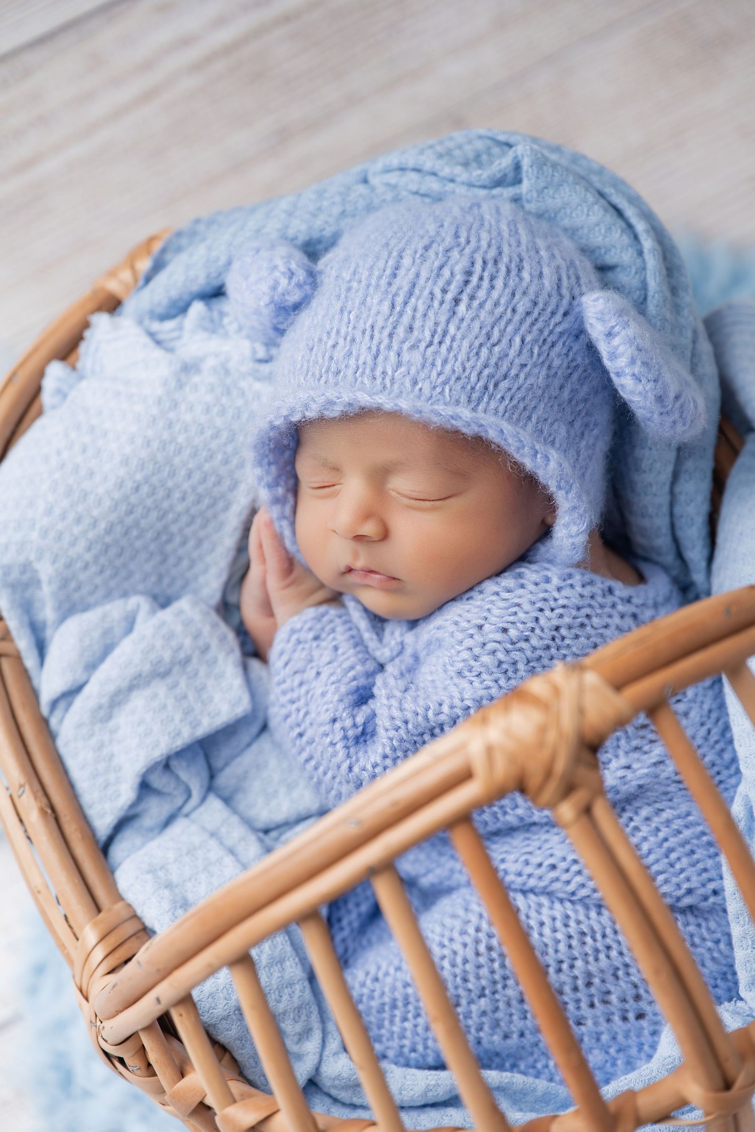 Sleeping newborn baby in blue knit hat with ears, wrapped in blue blanket inside a wicker bassinet.
