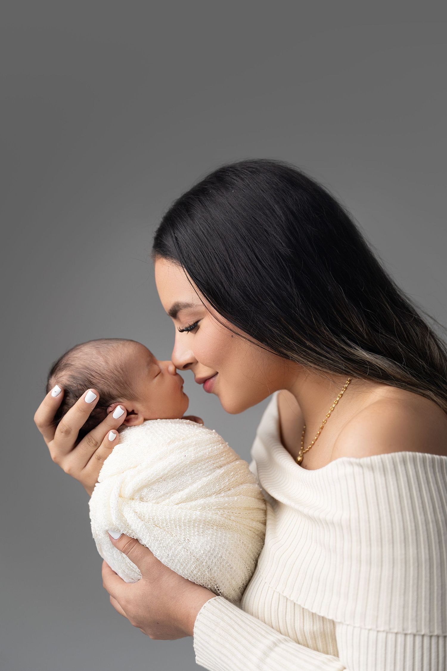 A smiling dark-haired mother in white holds her swaddled newborn baby close, touching noses tenderly on gray background.